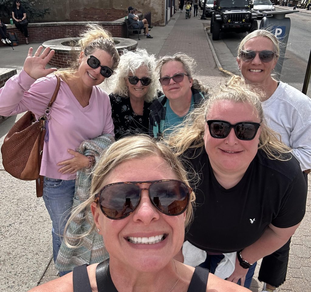 Group of women smiling and connecting during a wellness retreat, gathered outdoors in a scenic setting—capturing the power of community, support, and shared experience.